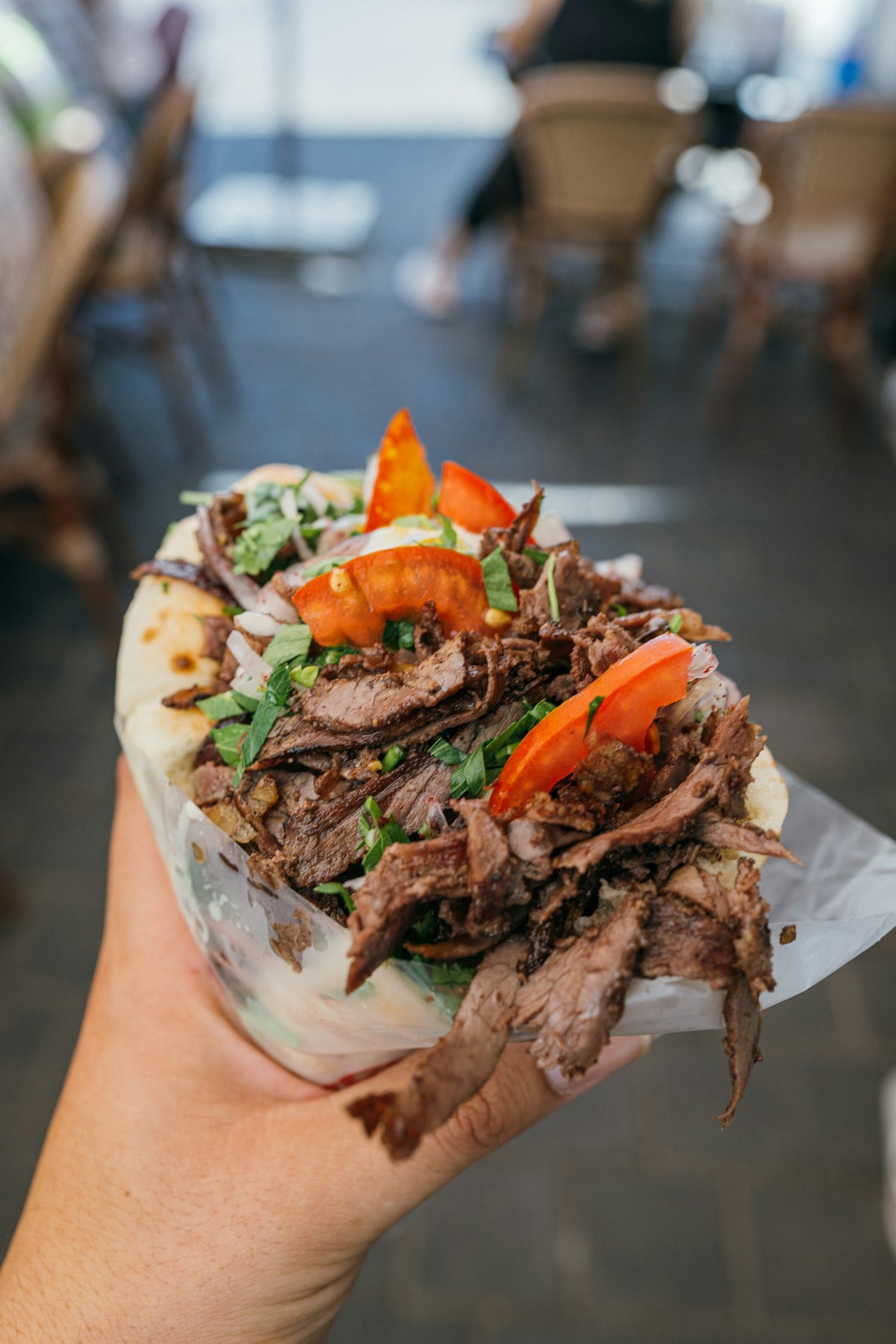 Hand holding a loaded gyro wrap with sliced meat, tomato, and herbs.